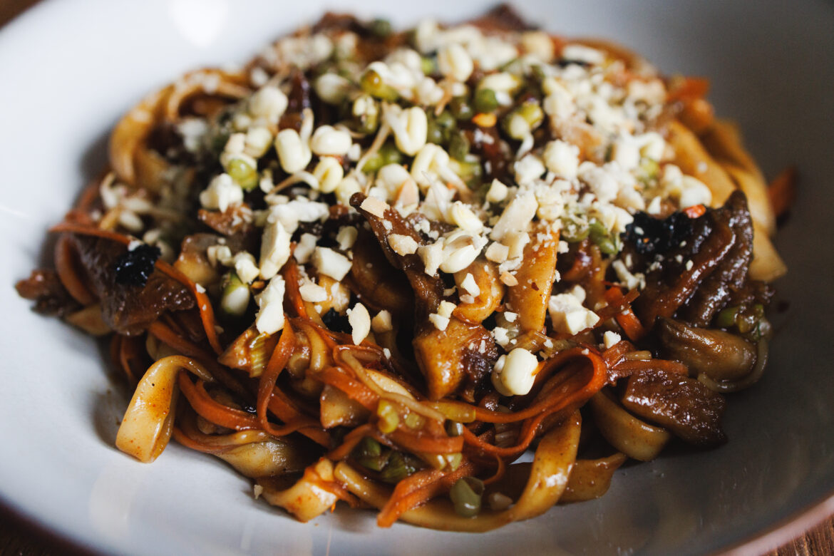 Veganské udon nudle s křupavou hlívou, žampiony, mrkví a tamarindovou omáčkou, sypané mungo klíčky, sekanými arašídy a chilli vločkami